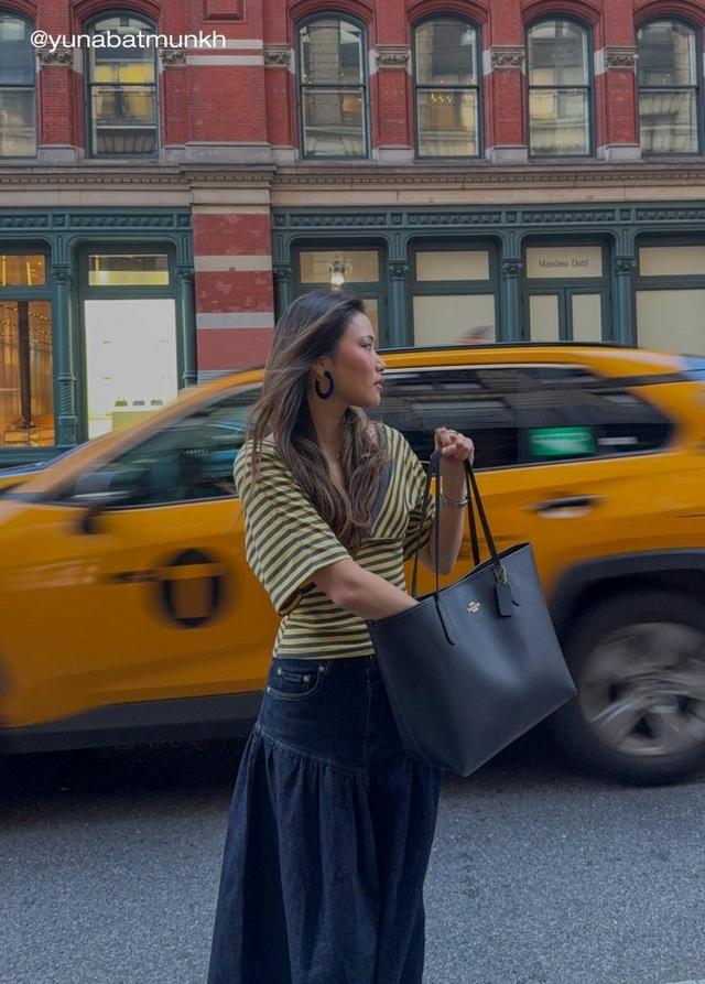 Woman wearing striped top and hoop earrings carries black leather tote bag on city street with taxi passing.