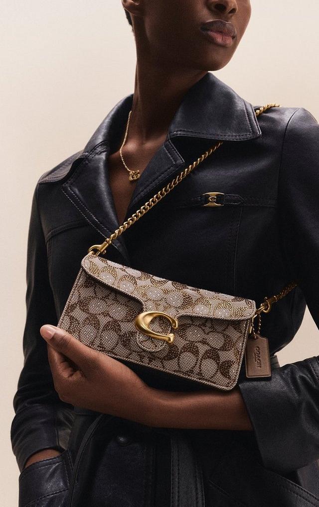 Model holding a Coach signature canvas crossbody bag with gold chain strap and hardware.