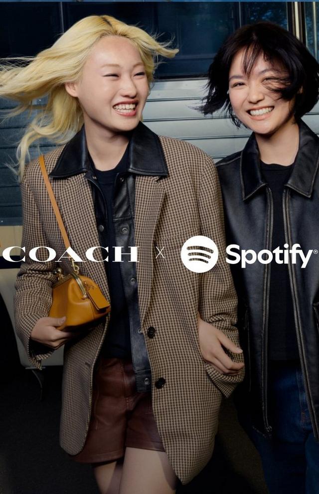 Two women laughing together carrying Coach handbags in front of a bus with COACH x Spotify TABBY TOUR text.
