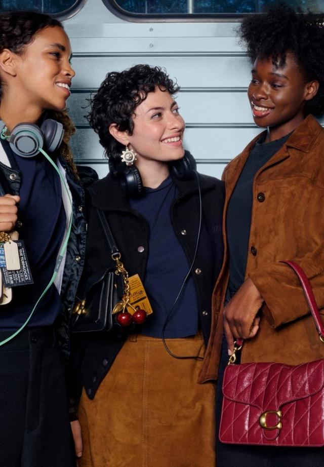 Trois femmes souriantes et discutant en portant des sacs Coach ornés de breloques, des vestes et des casques.