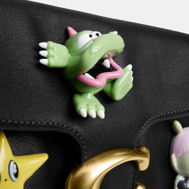 Green dinosaur charm with pink details and white claws attached to black leather bag.