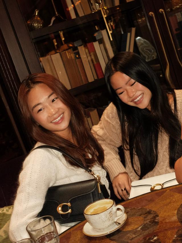 Three women smiling at a cafe table with drinks, handbags, and an open book.